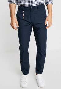 Uomo che indossa pantaloni blu scuro, sneakers bianche con bande in velcro, e una camicia a quadri con le maniche arrotolate.