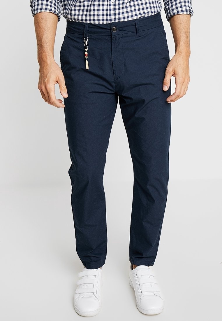 Uomo che indossa pantaloni blu scuro, sneakers bianche con bande in velcro, e una camicia a quadri con le maniche arrotolate.