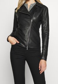 Svart läderbikerjacka med diagonalt blixtlås, halskrage och smal passform, som bärs ihop med en grå topp och svarta byxor.