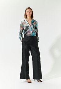 Camicia di seta a fantasia con tonalità di turchese, rosa e nero; abbinata a pantaloni a gamba larga neri a vita alta; semplici scarpe con cinturini.
