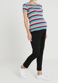 Zwangerschaps-T-shirt met horizontale strepen in turquoise, rood en blauw; gecombineerd met zwarte broek. Witte sneakers, minimalistisch design.