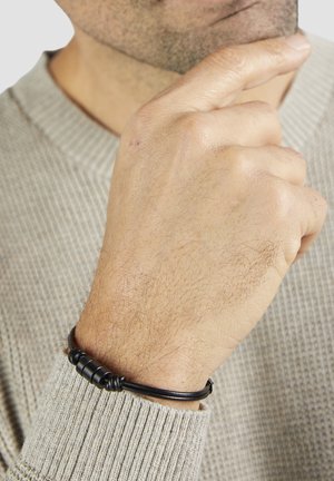 Schwarzes Lederarmband mit zylindrischen metallischen Akzenten, getragen an einer Hand mit heller Haut, vor einem strukturierten beigen Pullover.