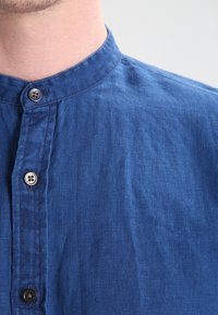 Modrá lněná košile s límcem bez klopy, se dvěma viditelnými knoflíky a texturovaným povrchem. Úhledné švy podél předního zapínání.
