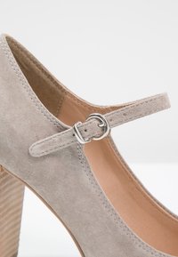 Chaussure à talon haut en daim beige avec une fine bride ajustable et une boucle argentée, mettant en valeur la texture et les coutures.