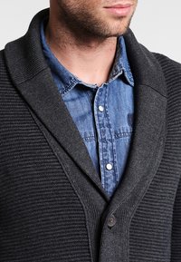 Mörkgrå stickad cardigan med en bred sjalkrage, med en enkel knappstängning över en ljusblå denimskjorta med synlig ficka.