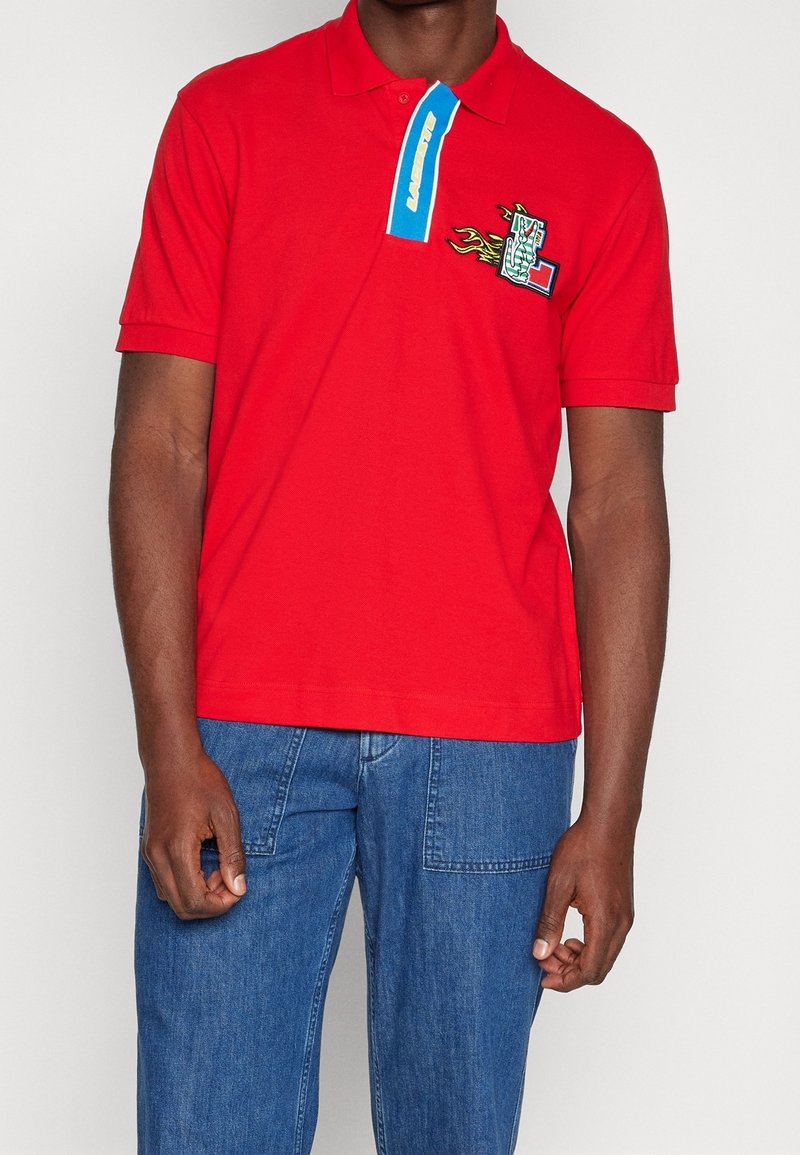 Homme portant un polo rouge avec une bande bleue et un écusson en forme de flamme sur la poitrine, associé à un jean bleu.