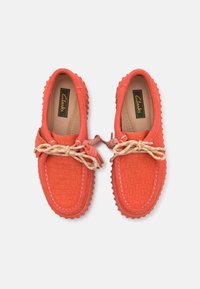 Mocasines rojos texturizados con un patrón trenzado, suela de goma y un lazo decorativo. Presentan cordones beige y el logo de Clarks en la plantilla.
