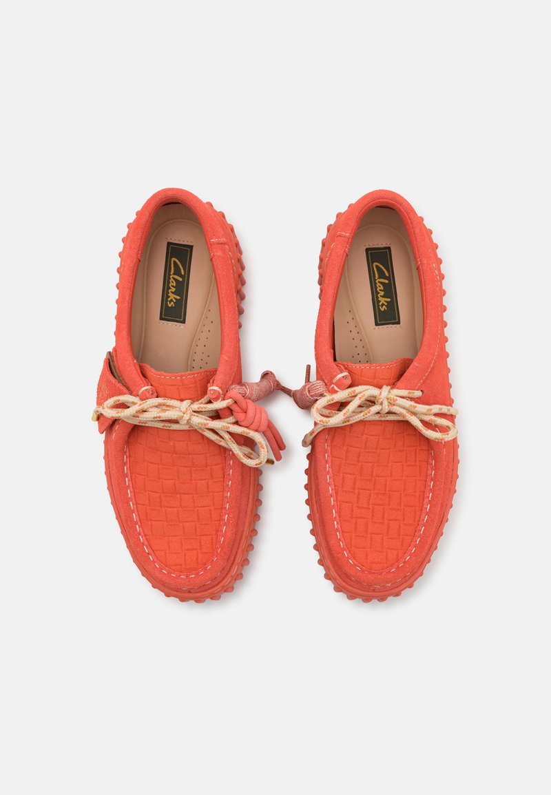 Mocassins rouges texturés avec un motif tissé, semelle en gomme et nœud décoratif. Présente des lacets beiges et le logo Clarks sur la semelle intérieure.