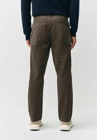 Eine Person trägt dunkelbraune Cordhosen und beige Sneaker und steht mit dem Rücken zu einer schlichten hellen Hintergrund.