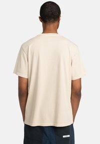 Bežová bavlnená tričko, krátke rukávy, okrúhly výstrih, s hladkou textúrou. Pohľad zozadu ukazuje jednoduchý, nezdobený dizajn.