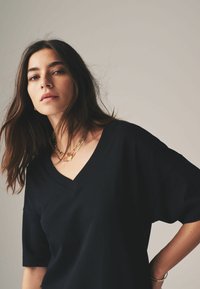 Ung kvinna med långt mörkt hår som bär en svart T-shirt med V-ringning och guldhalsband, poserande med ett neutralt uttryck mot en enfärgad bakgrund.