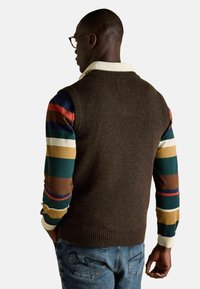 Pruun kudumvest mitmevärvilise triibulise pika varrukaga särgi peal. Vestil on ribakrae ja -äär. Mugavad sinised teksad.