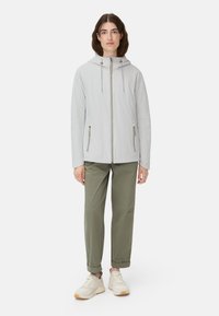 Hellgraue Jacke mit Kapuze, Frontreißverschluss und zwei Seitentaschen. Kombiniert mit olivgrünen Hosen und weißen Turnschuhen. Einfaches, schlankes Design.