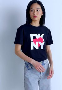 Svart bomull T-skjorte med "DKNY"-logo i hvitt og rødt. Har korte ermer og en avslappet passform, kombinert med lyseblå denimjeans.