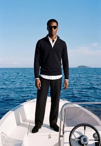 Homme portant un pull noir, une chemise blanche, un pantalon noir et des lunettes de soleil, debout à la proue d’un bateau-moteur blanc sur une eau bleue calme.