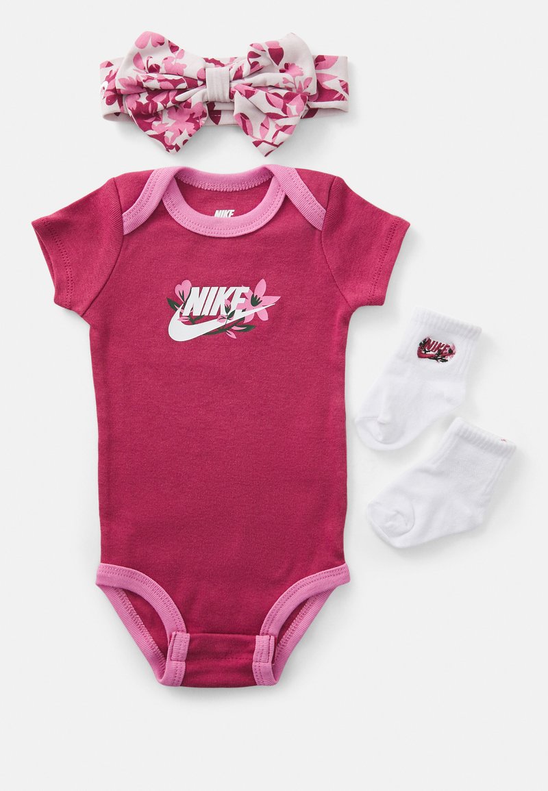 Macacão rosa de algodão com mangas curtas, com detalhes em rosa contrastante e o logo da Nike adornado com acentos florais. Inclui meias brancas com o logo e uma faixa de cabelo floral.