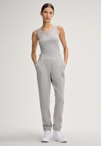 Grijze sportoutfit bestaande uit een mouwloos topje en joggers met elastische tailleband, met een subtiel logo detail. Draagt witte sneakers.
