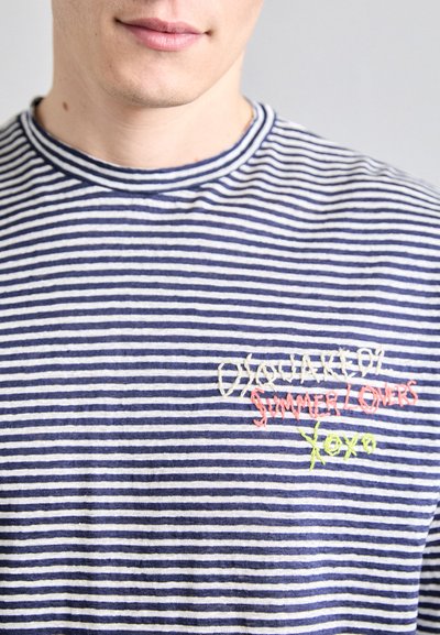 Tricou cu dungi în albastru și alb din bumbac, având guler rotund și text brodat în roz și verde pe partea stângă a pieptului.