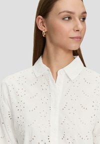 White cotton shirt with floral eyelet pattern, button-up front, and a classic collar, featuring a smooth texture and short sleeves.