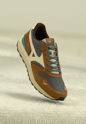Chaussure de course Mizuno marron, grise et crème avec lacets et semelle texturée, posée sur une surface douce de couleur claire.