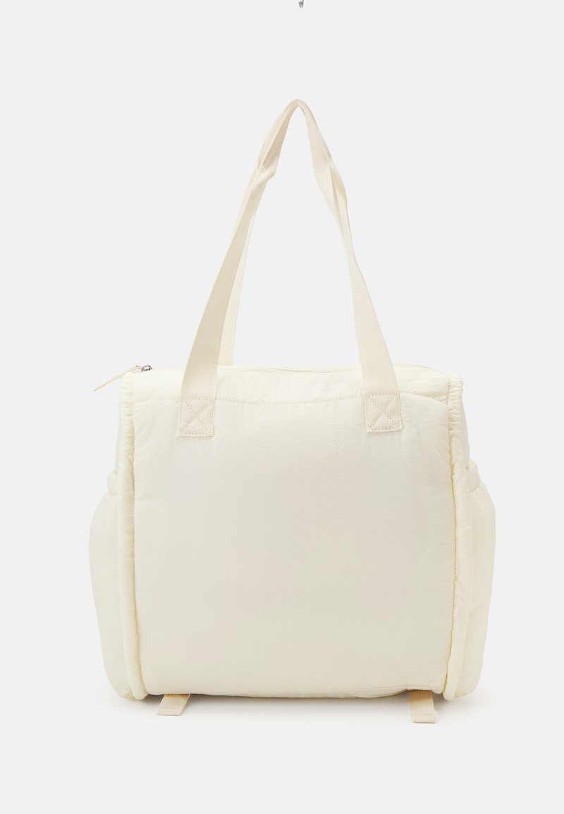 Sac fourre-tout de couleur crème avec une texture douce et matelassée et deux poignées solides. Il dispose d'une fermeture éclair et de poches latérales pour un rangement supplémentaire.