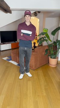 Maroon and navy long-sleeve polo, gray denim jeans, white sneakers. Indoor setting with a plant and corduroy sofa in the background.