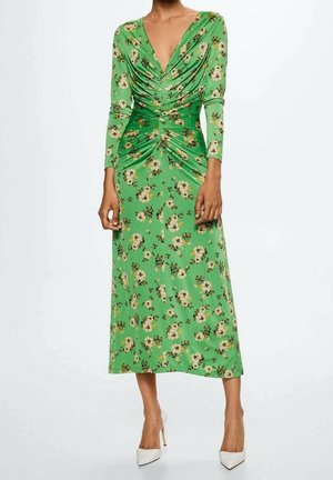 Frau trägt ein grünes Midikleid mit Blumenmuster, gerafften Details und tiefem V-Ausschnitt, dazu weiße High-Heels.