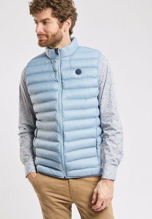 Gilet rembourré bleu clair avec un matelassage horizontal, fermeture éclair, col montant et patch logo circulaire. Porté par-dessus une chemise à manches longues à fleurs.