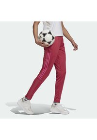 Czerwone spodnie sportowe z różowymi paskami, zwężanym krojem i elastycznymi mankietami. Model trzyma czarno-biały piłkę nożną, ubrany w białe sneakersy.