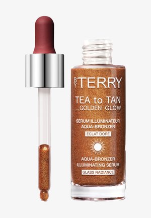Sérum bronzant liquide marron scintillant dans un flacon en verre transparent avec pipette et bouchon argenté étiqueté "By Terry Tea to Tan Golden Glow."