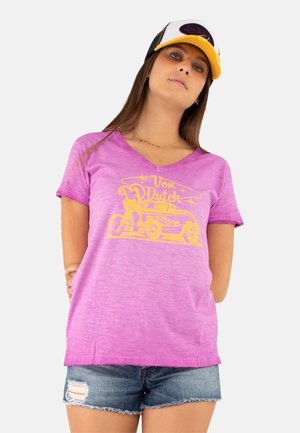 Femme portant un t-shirt rose Von Dutch, un short en jean bleu et une casquette de baseball jaune et blanche Von Dutch sur un fond uni.