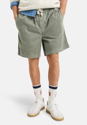 Shorts en coton verts avec une taille élastique et un cordon de serrage. Présente des poches latérales et une coupe décontractée. Associés à des baskets blanches et des chaussettes rayées.