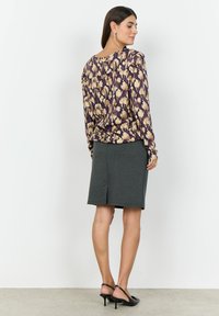 Blus med blommönster i lila och guld, kombinerad med en mörkgrå knälång kjol med slits bak. Modellen bär svarta sandaletter med klack.