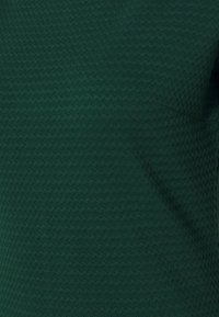 Mörkgrön strukturerad tyg med ett upprepande zigzagmönster, med en mjuk, slät finish och en lätt elastisk känsla.