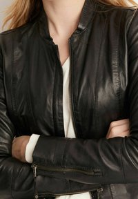 Kvinna med korslagda armar, bär en svart skinnjacka över en vit skjorta, visar jackans dragkedja och sömmar.