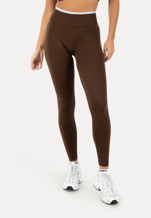 Kobieta w brązowych legginsach z wysokim stanem, połączonych z białymi sportowymi butami i skarpetkami do kostek na jednolitym jasnym tle.