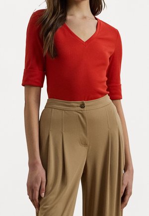 Woman wearing a red V-neck top with 3/4 sleeves tucked into high-waisted, pleated beige trousers with a button closure.