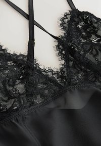 Haut de lingerie en satin noir avec une délicate bordure en dentelle florale et de fines bretelles réglables, sur un fond clair.