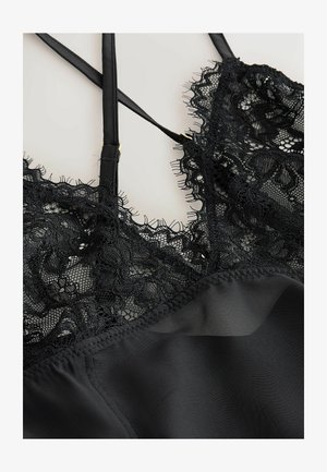 Melna satīna apakšveļas virskrekls ar smalku ziedu mežģīņu apdari un plānām regulējamām plecu siksnām uz gaiša fona.