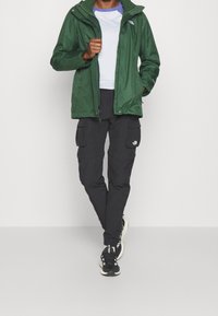 Giacca verde impermeabile con cappuccio, maglietta grigia a maniche lunghe, pantaloni cargo neri e sneakers nere con dettagli bianchi.