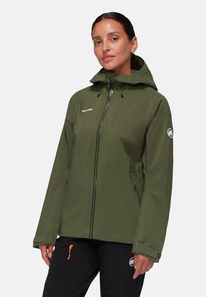 Frau trägt eine olivgrüne Kapuzenjacke mit Frontreißverschluss und schwarze Hose, steht vor einem schlichten hellen Hintergrund.
