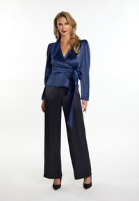 Top avvolgente in satin navy con maniche lunghe e vita legata; abbinato a pantaloni neri a gamba larga e tacchi neri. Tessuto morbido con un leggero lucido.