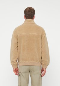 Casaco de pulôver em fleece bege com gola alta, punhos e bainha canelados. Exterior texturizado e forro suave. Sem hardware visível.