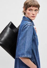 Πουκάμισο από τζιν σε ανοιχτό μπλε χρώμα με κοντά μανίκια, που διαθέτει κολάρο με κουμπιά και τσέπη στο στήθος· το μοντέλο κρατάει μια μαύρη δερμάτινη τσάντα tote.