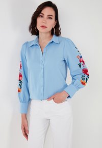 Light blue button-up shirt with puffed sleeves, featuring vibrant floral embroidery in red, green, and orange on the sleeves. White pants.