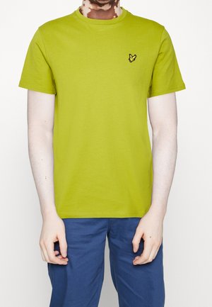 Person wearing a lime green short-sleeve t-shirt with a small embroidered yellow bird logo on the left chest and blue pants.
