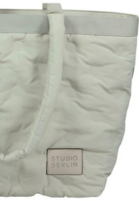 Quiltad ljusgrå tygväska med en texturerad yta, med ett mjukt handtag och en läderetikett som säger "STUDIO BERLIN."