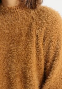 Maglione color ruggine realizzato in filato peloso e testurizzato con collo alto. Presenta una vestibilità ampia e dettagli a coste sul colletto.