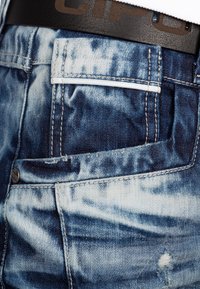 Jean en denim avec un dégradé de bleu clair à foncé, une texture délavée, un accent de couture blanche et une ceinture en cuir noir avec une lettrage en relief.