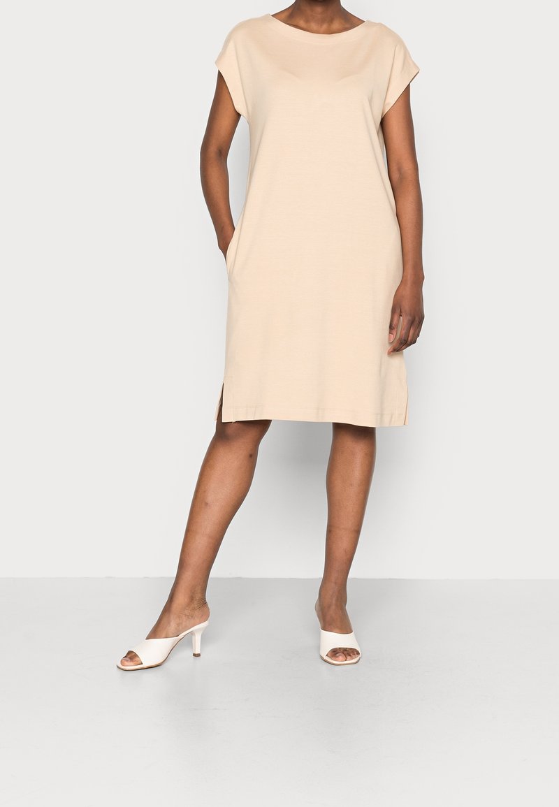 Béžové šaty s krátkými rukávy, volného střihu, s bočními kapsami a mírnými rozparky na spodním lemu. Hladká texturovaná látka a délka ke kolenům.